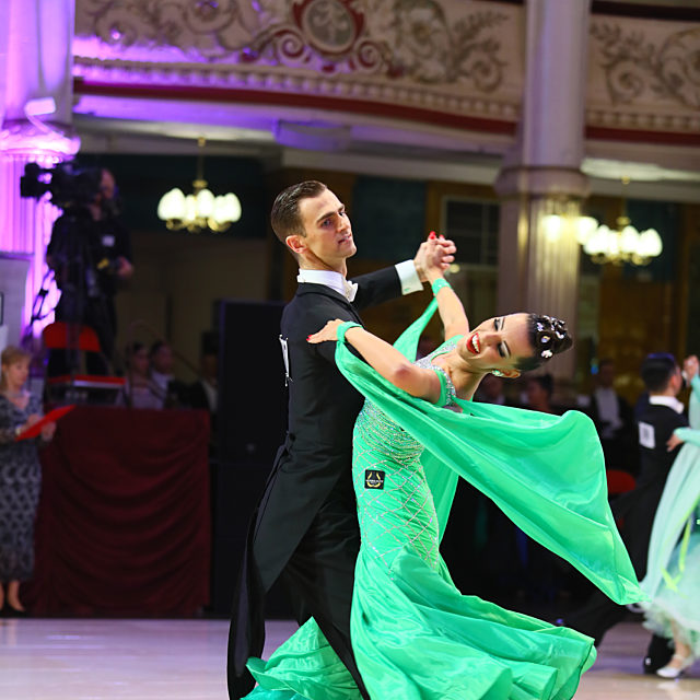 A pair of ballroom dancers dancing. She wears a green floating dress and he wears a suit.